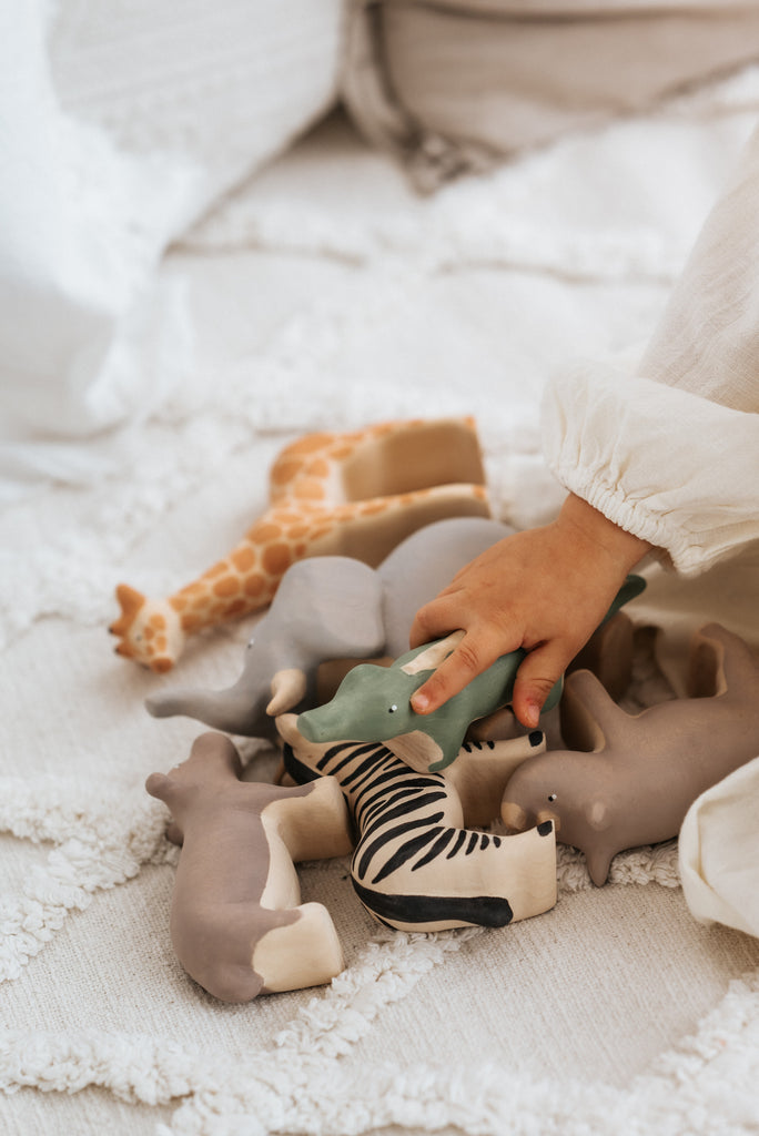 A child's hand reaching for a handcrafted linden wood Handmade Wooden Rhinoceros, painted with non-toxic paint by Meshka and Friends, on a textured white blanket. The collection includes a zebra, giraffe, elephant, and some other indistinguishable animals. The child is wearing a cream-colored long-sleeve shirt.