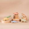 A collection of pastel-colored toy dinosaurs is arranged on wooden pedestals against a beige background. Handcrafted wooden toys like the Handmade Wooded Stegosaurus Dinosaur, along with a triceratops, parasaurolophus, brachiosaurus, and other species, feature non-toxic paint for safe play.