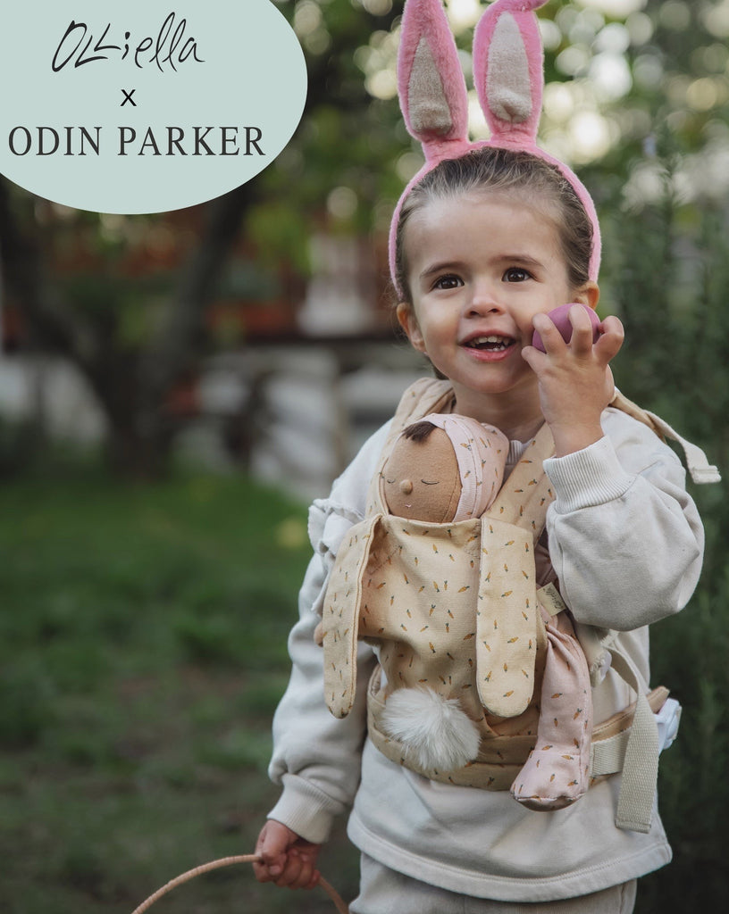 A joyful toddler girl wearing bunny ears and holding up an apple, with a Olli Ella x Odin Parker Dinkum Dolls Cottontail Carrier – Hopscotch around her chest, with the logo "Olli Ella x Odin Parker" at the top