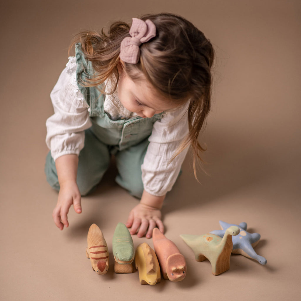 A young child plays with a handmade Wooden Parasaurolophus Dinosaur, coated in child-safe paint, on the floor, focusing intently on arranging them in a line.