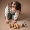 A young child with long hair, dressed in a white top and green overalls with a pink bow in their hair, kneels on a beige surface, arranging colorful handcrafted wooden toys. Their attention is focused on the Handmade Wooden Stegosaurus Dinosaur and other dinosaurs lined up in front of them.