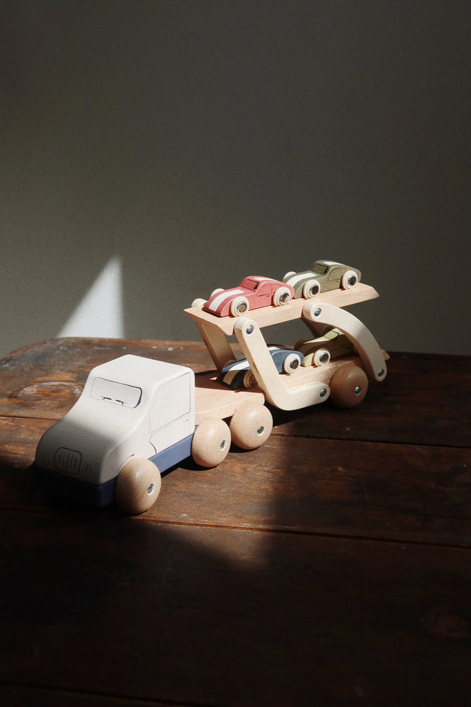 The Konges Sløjd - Wooden Car Transporter, made from FSC-certified beech wood, carries three small race cars on a rustic wooden surface. Sunlight casts a narrow beam of light across the scene, highlighting the toys against a muted background.