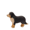 A handcrafted wooden figurine of a dog painted with black, brown, and beige colors. The dog is standing on all four legs with its head facing left. Made from sustainable materials, this piece exemplifies the quality of the Ostheimer Bernese Mountain Dog, set against a plain white background.