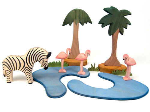 A wooden toy scene featuring Ostheimer toys: a zebra, three flamingos, two palm trees, and Ostheimer Pond Plates - Set of 2 representing water. With green foliage meticulously added, the elements seem arranged to depict a miniature oasis or wildlife habitat perfect for imaginative play.