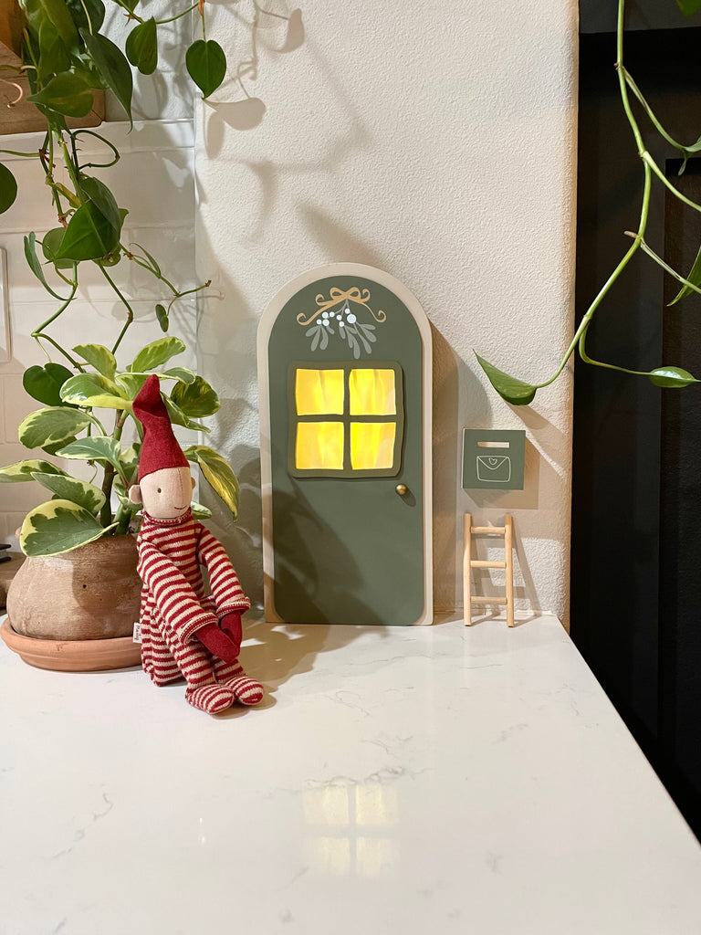 A small decorative door with a yellow glowing window is mounted on a wall in a pixy style. A tiny ladder leans against the wall near the door. A Maileg Long Legged Pixy wearing a red and white striped knitted suit and hat sits beside a potted plant with trailing vines.