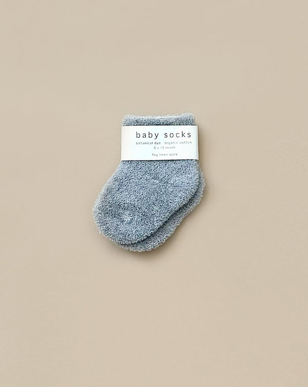 A pair of soft, blue baby socks crafted with pile knitting features a label that reads "Baby Pile Knit Socks - Blue" and includes additional details. The fluffy socks are displayed on a neutral beige background.