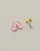 Placed on a light beige background, a pair of Minikane Beach Sandals in Petal Pink—a small woven beach accessory for babies—rests next to a sprig of yellow billy button flowers tied together with string. Perfect for tiny feet, these sandals evoke the charm of Minikane dolls and sunny days by the sea.