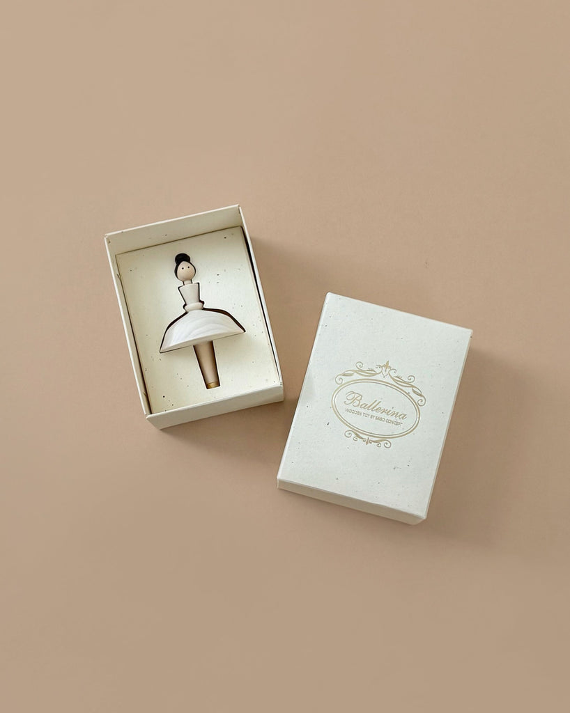 A small, white rectangular box featuring a delicate illustration of a ballerina inside, partially opened on a beige surface. The hand-painted lid of the Wooden Ballerina box is adorned with an elegant border and the word "Ballerina," crafted with meticulous detail using water-based paint.