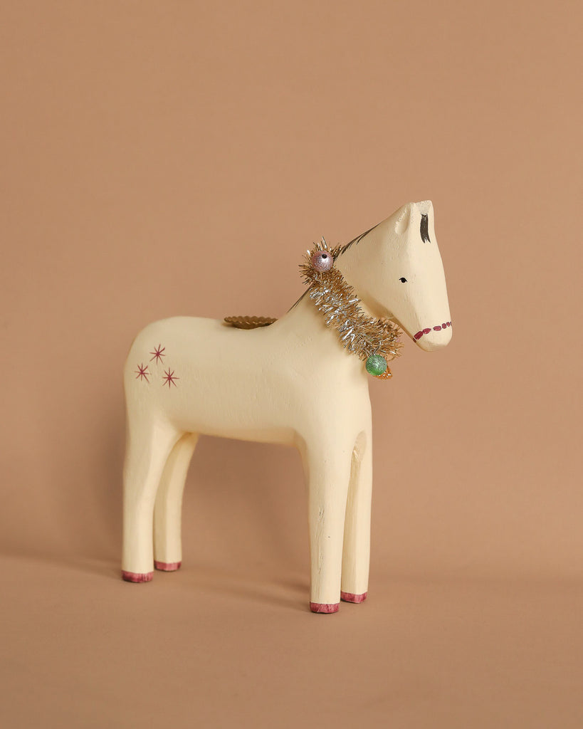 The Maileg Wooden Horse Candle Holder - Small, a handcrafted cream-colored figurine, features pink stars and red-tipped hooves. It stands against a beige background and is adorned with a decorative collar embellished with silver tinsel and small ornaments.