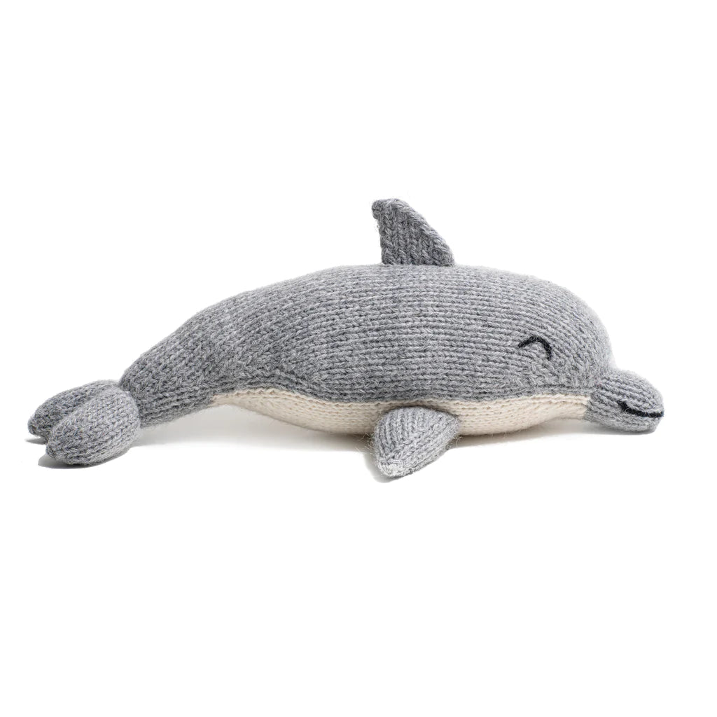 A Knit Alpaca Dolphin toy, handcrafted in Peru, is shown resting on a white surface with a smiling face and small fins.