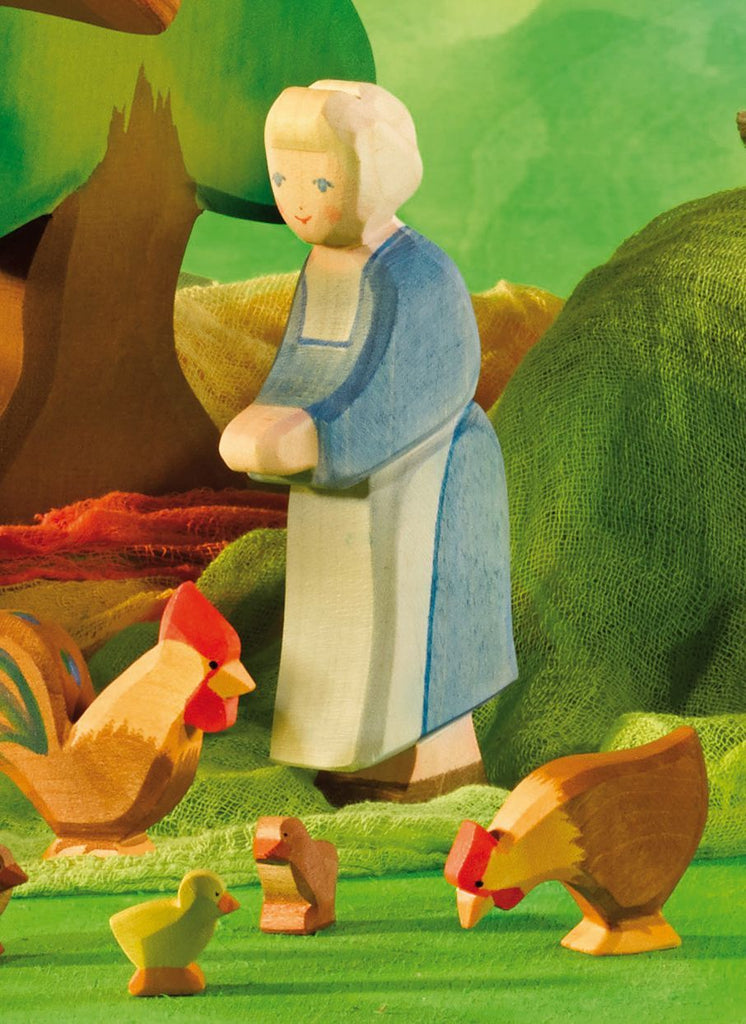 The Ostheimer Grandmother, a wooden figurine with light hair and dressed in a blue and white outfit, stands among handcrafted wooden chickens and chicks on a green landscape. A tree and textured green backdrop complete the scene, making it ideal for imaginative play with Ostheimer toys.