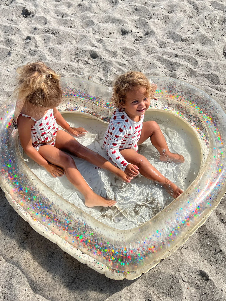 Two young children sit in an Inflatable Heart Pool filled with water on a sandy beach. Both kids are wearing white swimsuits with red patterns and are smiling while playing in the shallow water under the bright sun, surrounded by glitter and confetti reflections from their durable PVC pool.