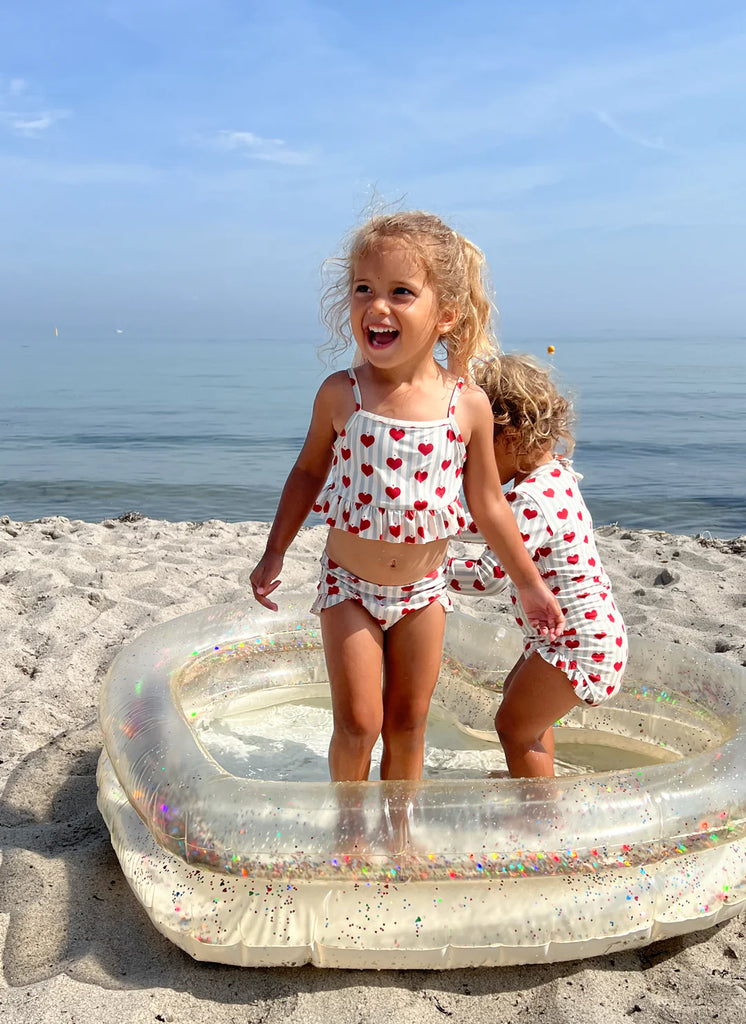 Two young children play in a small Inflatable Heart Pool on a sandy beach. One child is standing up and laughing, while the other, slightly crouched and facing away, appears to be playing with the water. Both wear coordinating swimsuits with red heart patterns. The ocean is visible in the background under a clear blue sky.