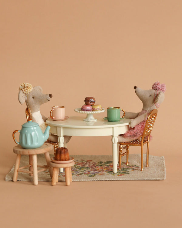 The Maileg Tea Party Starter Set features two plush ballerina mice dolls at a small table adorned with diverse pastries and vibrant mugs. A teal teapot and a bundt cake are placed on adjacent stools, creating a cozy scene where one mouse sits gracefully in an elegant gold chair, all set against a warm brown backdrop.