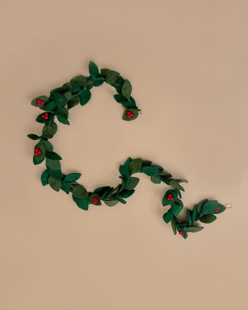 Introducing the 6' Felt Holly Garland: a beautifully handcrafted piece featuring green wool felt leaves adorned with small red flowers, artfully arranged on a beige surface in a serpentine design. Perfect for holiday decor, it brings a charming and delightful touch to any setting.