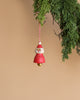 A Wooden Santa Ornament is suspended from a branch of greenery, featuring a red and white design with a cheerful face and a small gold bell inside, all set against the backdrop of natural wood.