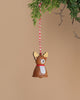 The Wooden Reindeer Ornament is an adorable decoration made from natural wood, complete with antlers and a red scarf. It hangs from a red and white striped string, containing a bell that sways gently from a green branch against a beige background.