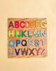 The Grimm's Alphabet Puzzle features a sturdy wooden board with a robust frame that displays colorful, non-toxic three-dimensional letter cutouts of the alphabet arranged in four rows. Each letter is painted in a different color, creating a vibrant and educational visual on a pale yellow background.