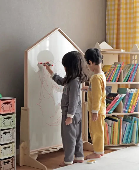 Two children in pajamas use the NORITER Signature Double-Sided Board Bundle to draw with magnetic alphabets in a cozy room. The space, adorned with pastel-toned crates and a bookcase filled with vibrant books, is made even warmer by a plaid curtain in the background.