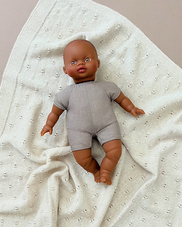 A Minikane Soft Body Doll (11") - Ondine with brown skin and phthalate-free vinyl, dressed in a simple gray outfit, lies on a textured white knitted blanket. The bald baby doll's arms and legs are slightly spread, and it has a peaceful expression—perfect as a bedtime companion.