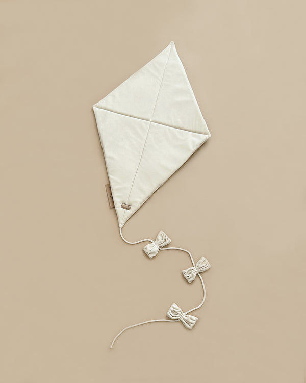 The Velvet Kite Wall Decor - Cream is a charming beige, diamond-shaped VELVET kite featuring two intersecting seam lines and a small tag near the bottom corner. Adorned with three decorative bows on its tail extending from the bottom point, this OekoTex®Standard100 certified piece makes a delightful addition to any children's room decor, all set against a light brown background.