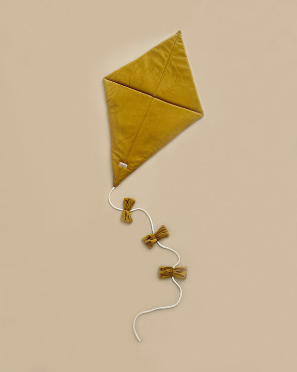 The Velvet Kite Wall Decor - Mustard, a mustard yellow, diamond-shaped kite, lies flat against a beige background. It features a white string with two matching yellow bows attached, making it an adorable piece for children's room decor.