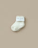 A single Baby Pile Knit Sock in natural white, fluffy and soft, with a label that reads "baby socks 0-12 months," is displayed against a plain beige background.