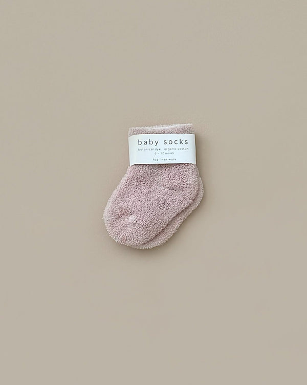 A pair of Baby Pile Knit Socks in pale pink is displayed against a neutral beige background. The fluffy, small socks are neatly folded, with a white label reading "baby socks" and "Newborn" visible on top.