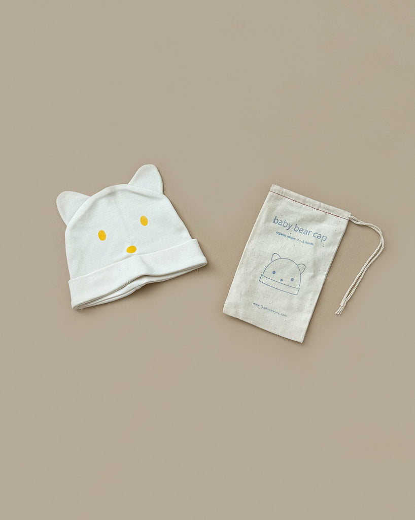 A light-colored organic cotton Baby Bear Cap, featuring adorable bear ears and a simple face design, lies on a beige surface next to a drawstring pouch. The pouch is labeled "Baby Bear Cap" and showcases an illustration of the hat, making it an ideal newborn gift.