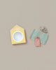 A minimalist setup on a light beige background features a small cardboard cutout resembling a beach cabin with an open circular window, beach accessories including a green and white striped mat, a tiny pink bag, and a colorful tiny object from the Maileg Beach Set for Big Sister.