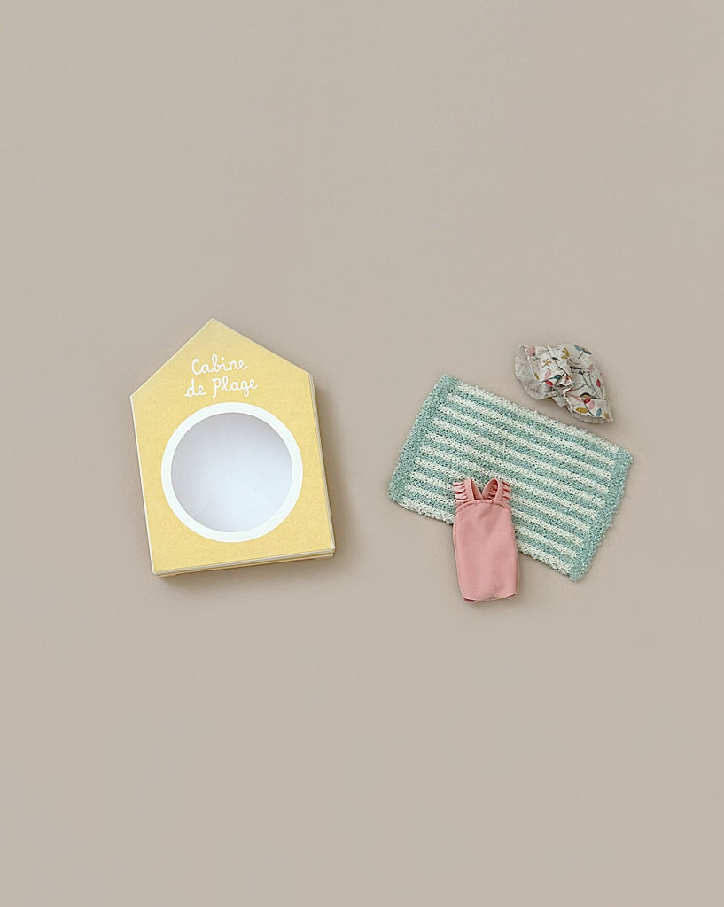 A minimalist setup on a light beige background features a small cardboard cutout resembling a beach cabin with an open circular window, beach accessories including a green and white striped mat, a tiny pink bag, and a colorful tiny object from the Maileg Beach Set for Big Sister.