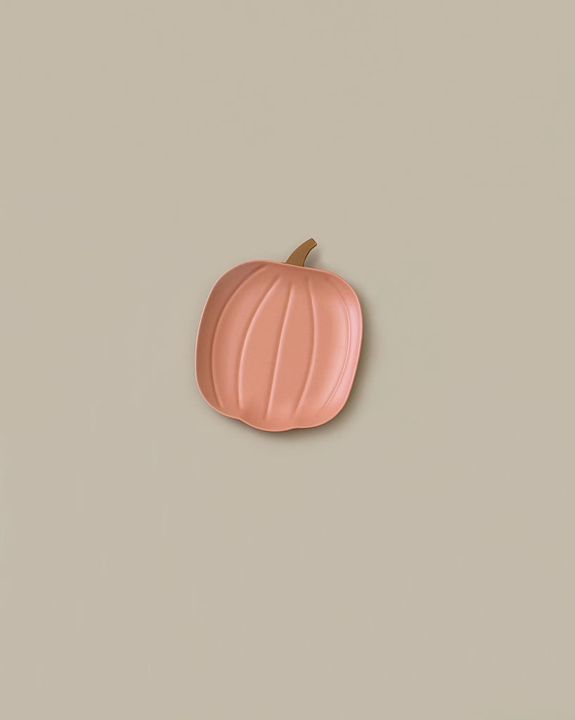 A Meri Meri Reusable Bamboo Pumpkin Plate, shaped like a minimalist pink pumpkin with a stem, centered against a beige background, creates an elegant Halloween plate display.