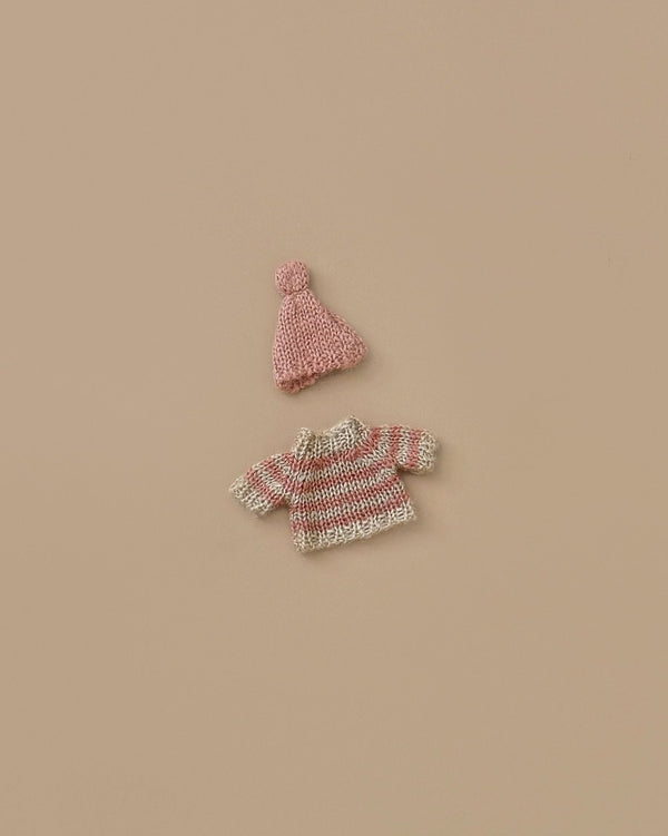 A small Maileg Knitted sweater and Hat for Big Sister Mouse in shades of pink and beige are arranged on a plain beige background. Ideal for winter, the hat sports a cute pom-pom on top—a snug outfit ready to be embraced by its big sister or brother.