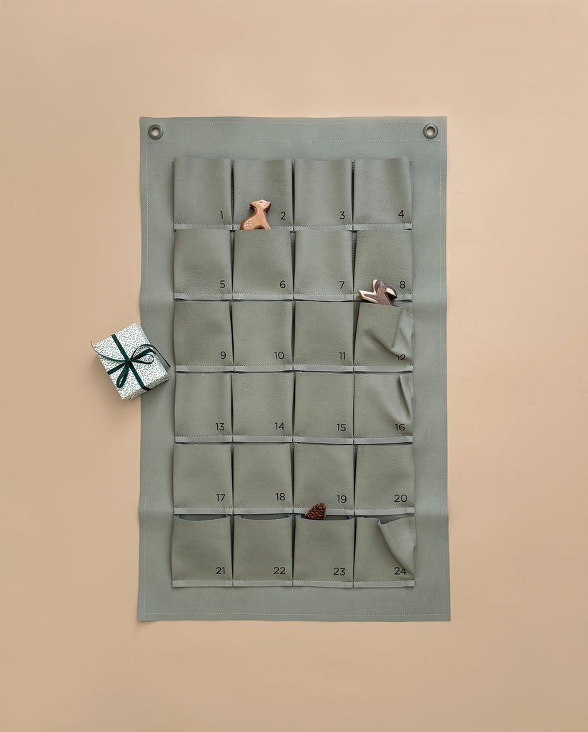 The Gathre Small Advent Calendar - Grove, featuring 24 numbered gift pockets in a minimalist fabric design, is displayed on a beige wall for an ideal Christmas countdown. Three of its pockets contain charming small toys: a cloth bunny, a peg doll, and a fabric pinecone. Next to it lies a petite silver gift box wrapped with a black ribbon.