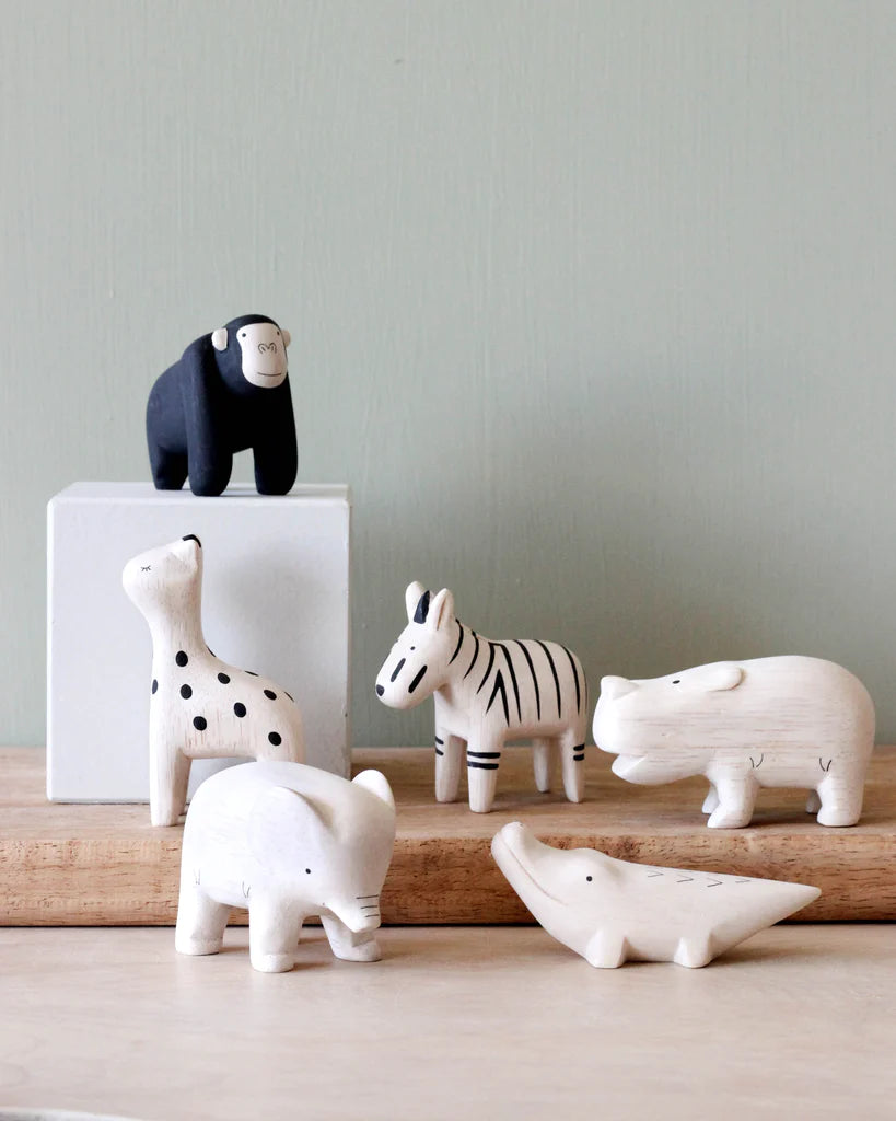 A set of white, handcrafted wooden animals, including a giraffe, zebra, rhinoceros, Handmade Tiny Wooden Safari Animals - Elephant, and crocodile, are arranged on a wooden surface. A black chimpanzee figurine stands on a white block behind them. The background is a light green wall.