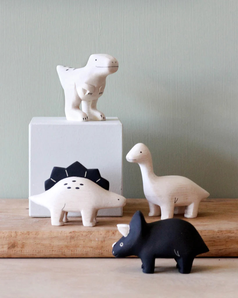 Four Handmade Tiny Wooden Dinosaurs - Stegosaurus on a wooden surface against a pale green background; includes a white t-rex on a pedestal, a white diplodocus, a white stegosaurus