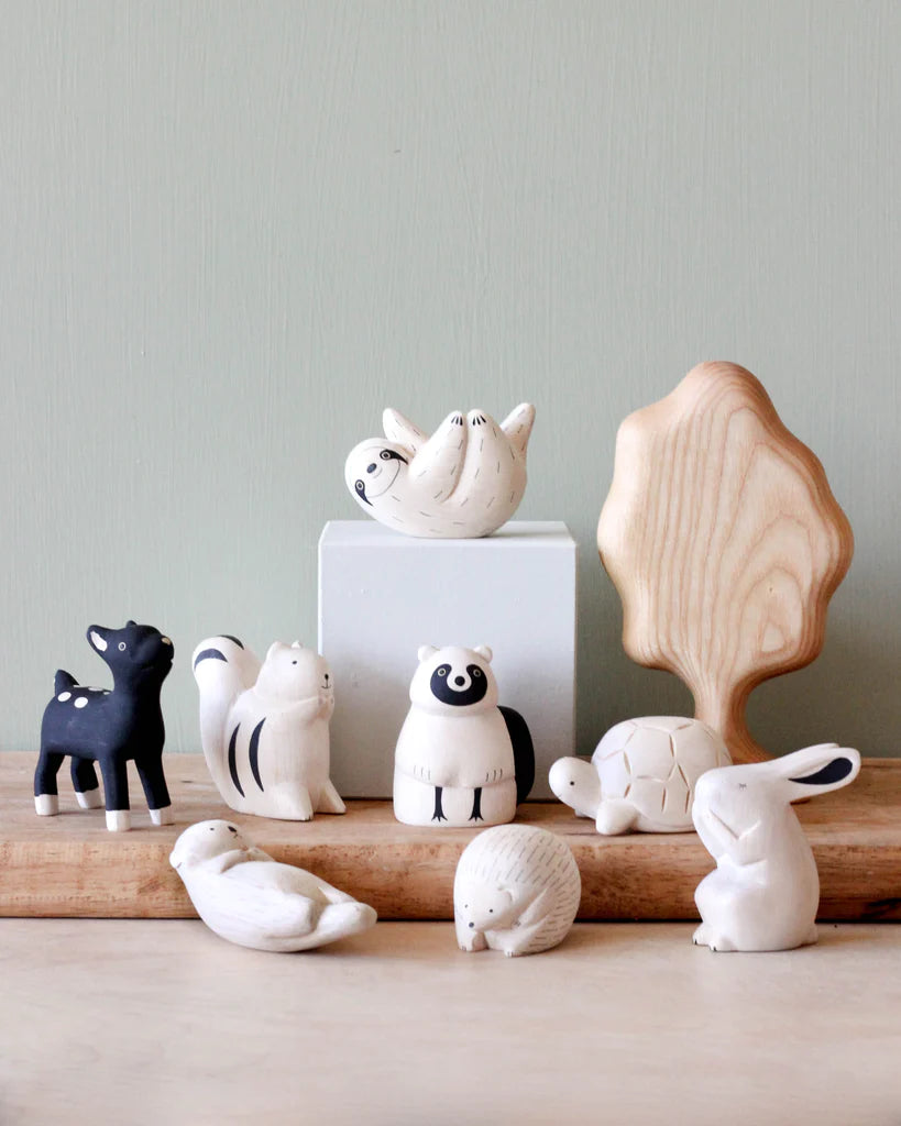 Displayed on a table is a charming collection of tiny, handcrafted forest animals made from Albizia wood and clay. This delightful group features a black deer, white squirrel, white raccoon, turtle, white rabbit, resting dove, and hedgehog. A wooden tree figurine stands behind them against a plain light green background—highlighting the exquisite Handmade Tiny Wooden Forest Animals collection.