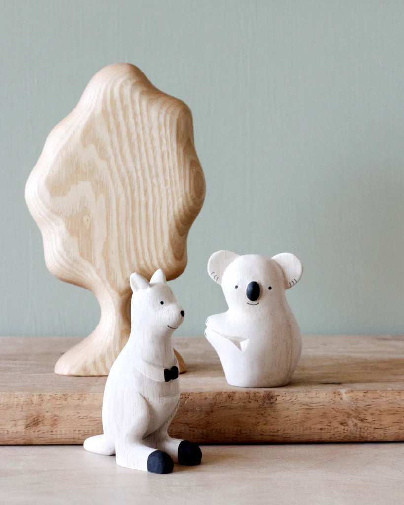 A Handmade Wooden Koala, crafted from Albizia wood, sits behind a charming handcrafted wooden toy kangaroo adorned with a black bow tie and black-tipped feet. They are placed in front of a large wooden tree on a wooden surface against a pale green background.