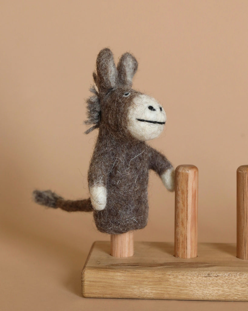 The Donkey Finger Puppet, handmade in Nepal from dark brown and white wool, stands smiling on a wooden peg in front of a beige background. This needle-felted donkey puppet features a bushy tail and is part of a wooden stand with two other empty pegs.