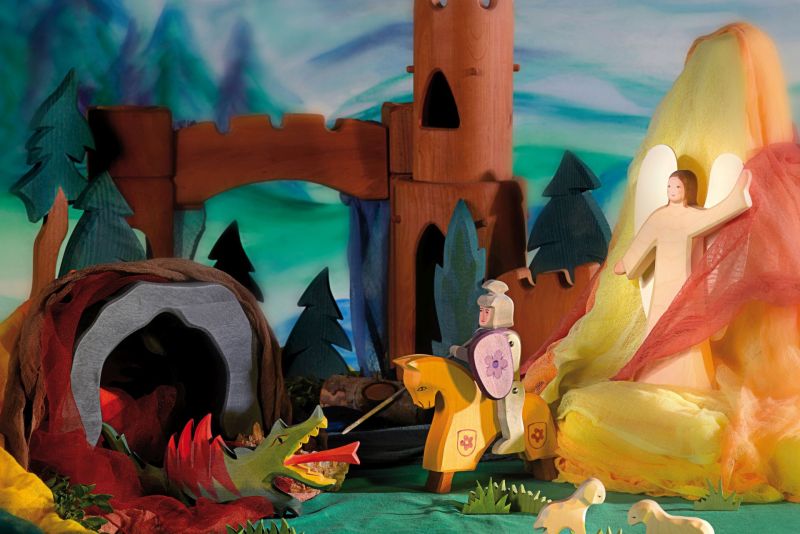 An intricately crafted scene features an Ostheimer Red Knight riding a yellow horse, bravely facing a green dragon emerging from a cave. Behind the knight, an angelic figure stands in front of a glowing yellow and orange backdrop. In the background, wooden castle towers and trees elevate this imaginative play with Ostheimer toys.