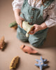 A child in overalls and a white blouse plays with a variety of handcrafted toys made from linden wood, including the Handmade Wooden Triceratops Dinosaur figure, while holding a small jug from which a white substance pours out.