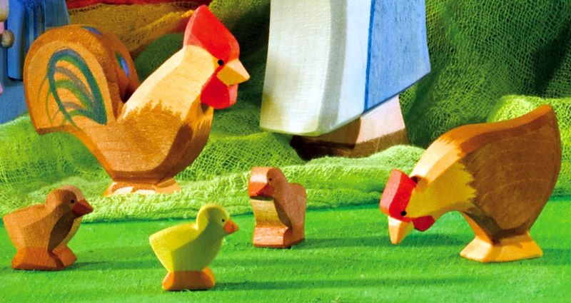 A close-up shot of Ostheimer brand handcrafted wooden farm animal figurines, including the Ostheimer Chick, Brown, on a green surface designed for imaginative play. The wooden figurines feature a rooster, a hen, and three chicks. In the background, a blue and white object is partially visible. The scene is set on textured green fabric.