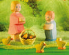 Ostheimer wooden figurines of a woman in a pink dress and a child in a blue outfit stand on a green woven surface. A small basket with flowers and grass is between them, and three Ostheimer Chick, Yellow are nearby. The background features textured green fabric and small plants, perfect for imaginative play.