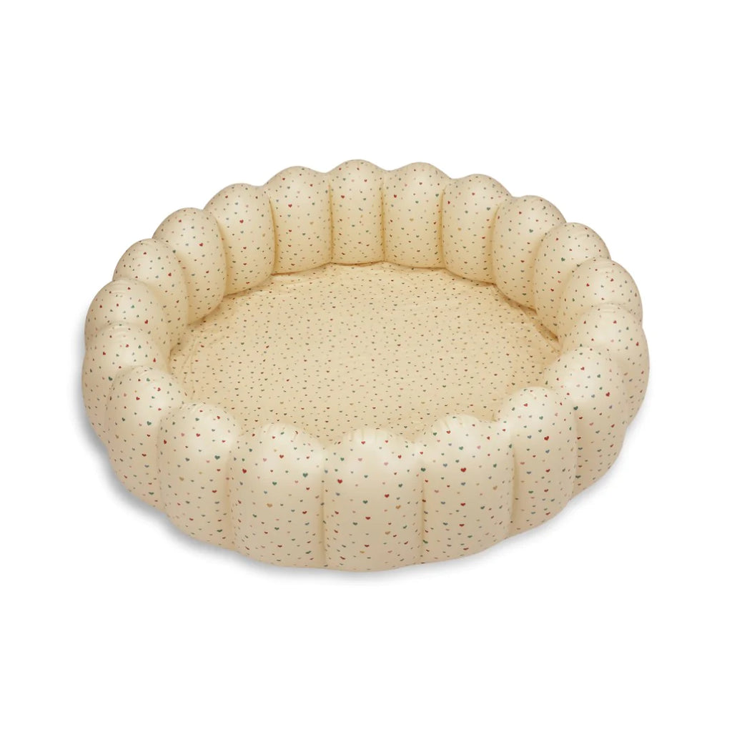 A round, beige pet bed with a quilted, doughnut-like design and subtle multicolored speckles. The edges are puffy and form a scalloped pattern, much like the gentle curves of an Inflatable Large Shell Pool - Multi Hearts, providing a cozy and cushioned area for pets to rest or sleep.