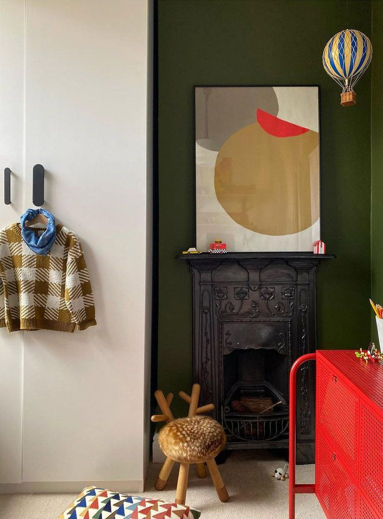 A cozy room features a green wall with a vintage black fireplace, adorned by a minimalist art piece. A colorful play area with toys and Faux Bambi Chairs enhances the playful ambiance.