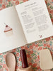 An open recipe book displaying a "cherry cupcake" recipe with an illustration of a cupcake on the left page and ingredients and directions on the right. Below the book, three wooden kitchen utensils rest on a floral-patterned surface, perfect for tiny bakers in training using the Madeline Baking Set - Petite Cousine.