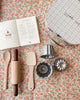 A flat lay photo of baking items on a floral patterned surface. There’s a cookbook opened to a cherry cupcake recipe, a rolling pin, a wooden spoon, a spatula, three metal baking molds, and the Madeline Baking Set - Petite Cousine—a perfect setup for tiny bakers just starting their kitchen adventures.