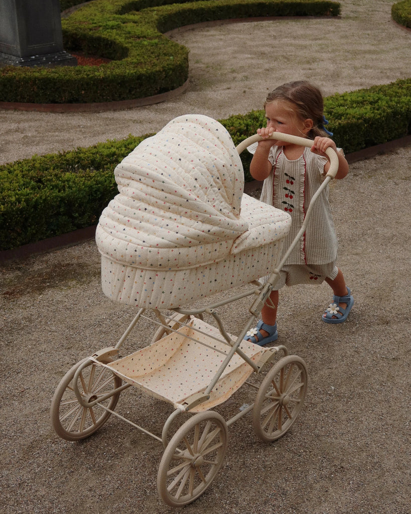 A young child in a light-colored dress pushes the Doll Pram - Multi Star with a closed canopy along a gravel path, set against neatly trimmed hedges in a garden setting.