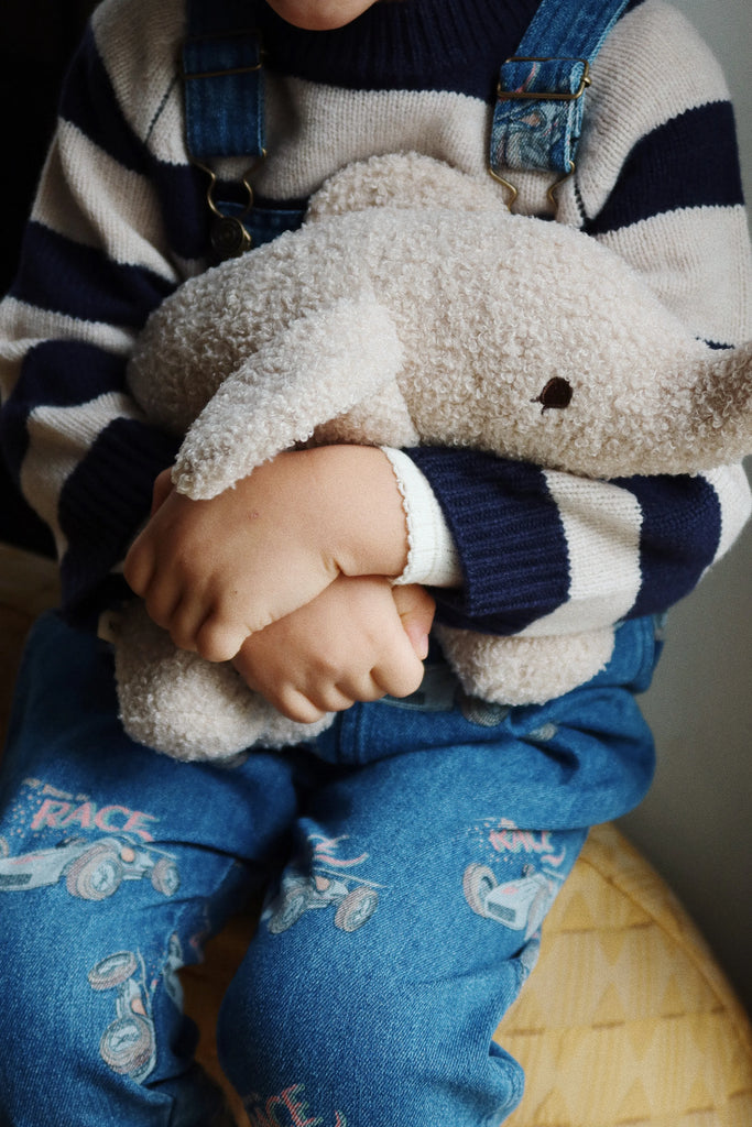 A child wearing blue overalls adorned with colorful patterns and a striped sweater cradles the Elephant Teddy Stuffed Animal with polyester filling in their lap, hugging it tightly. The child's face is not visible in the image.