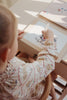 A child is drawing on a piece of paper in a spiral-bound sketchbook using a white crayon. The child is sitting at a light-colored desk and wearing a patterned, frilly outfit. The drawing features colors and shapes, but the specific details are not clear. Nearby lies My Magic Water Book - Off White, ready for more artistic fun.